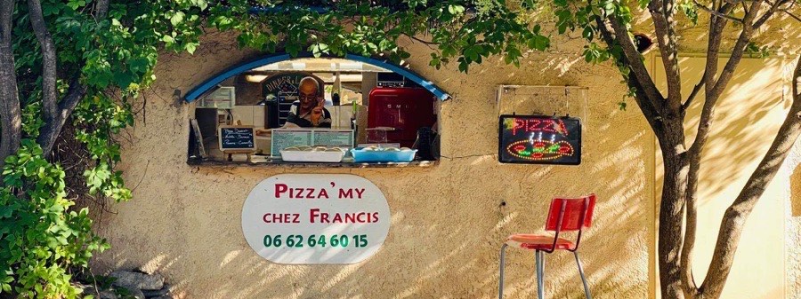 La devanture de Pizza'My : une fenêtre voûtée dans un mur ocre sous les figuiers, l'enseigne « Pizza'My chez Francis », et Francis à son comptoir